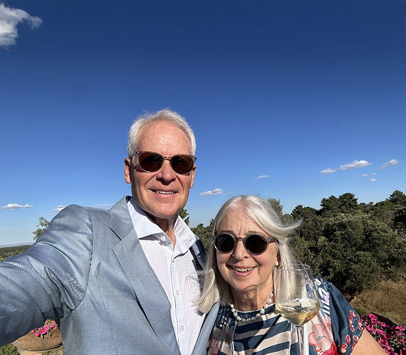 Alison and Craig Marthinsen, owners of Beachcomber villa and Blue Lagoon villa, Virgin Gorda, smiling together arm-in-arm with green trees and a bright blue sky in the background.