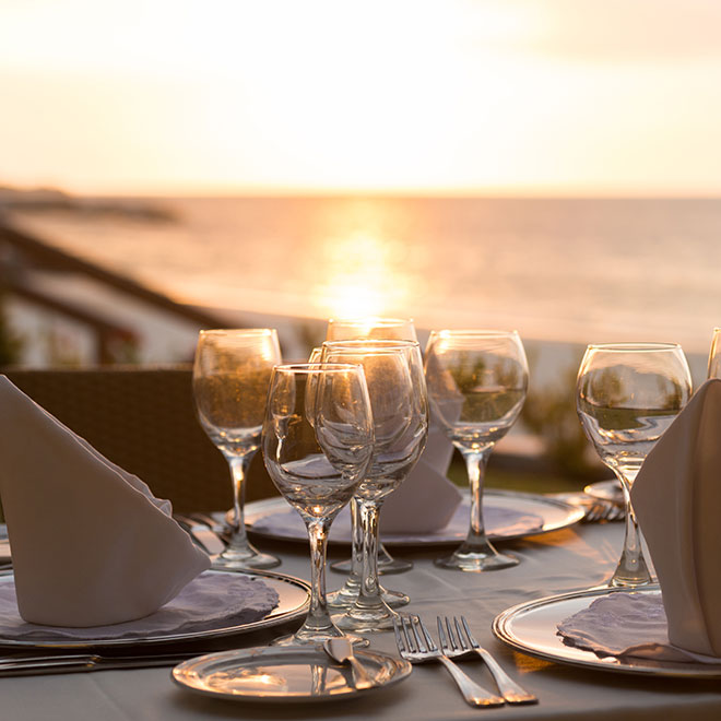 A dinner table set with classes, linens and silverware with a villa and the setting sun shining through in the slightly blurred background.