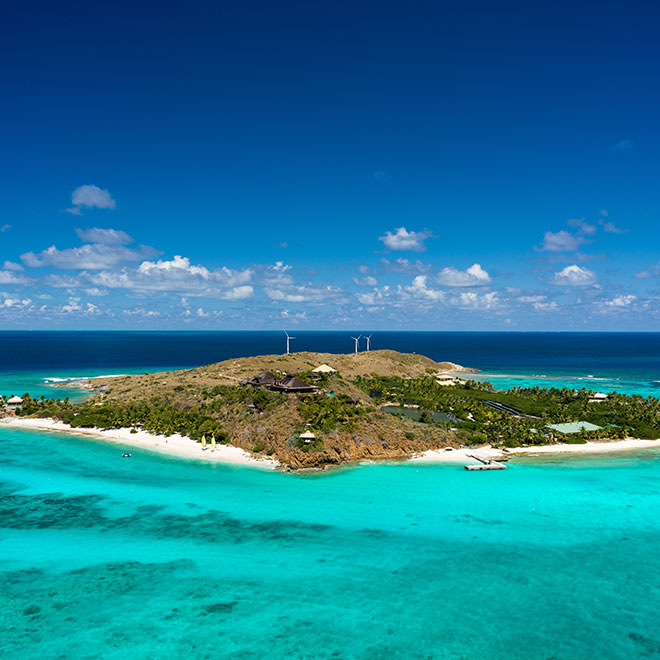 Necker Island, a tiny resort island surrounded by crystal blue water and coral reefs on a bright  sunny day with a cloud-dotted sky in the background.