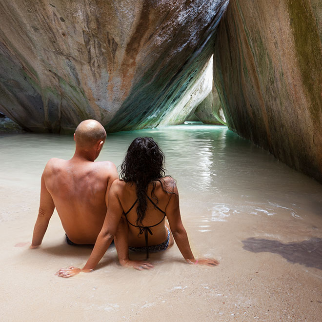 A couple sitting close together on the sand looking out on the boulder formations  of The Baths, Virgin Gorda, with the sun piercing through between the rocks.
