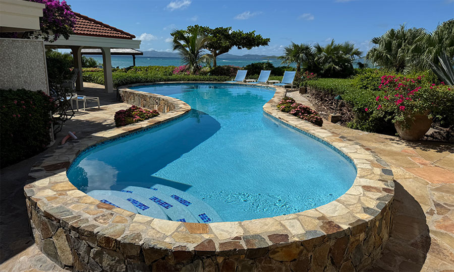 Beachcomber villa swimming pool surrounded by a stone patio and lush tropical garden with Mahoe Bay and blue sky in the background.