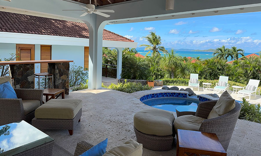 Blue Lagoon villa covered patio with comfortable seating looking out on the villa’s swimming pool with palm trees and the ocean in the distance.