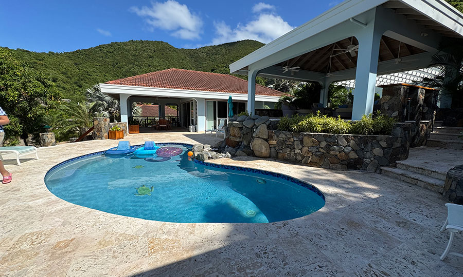 Blue Lagoon villa swimming pool bordered by a light stone sun deck adjacent to a large covered patio with the villa in the background.